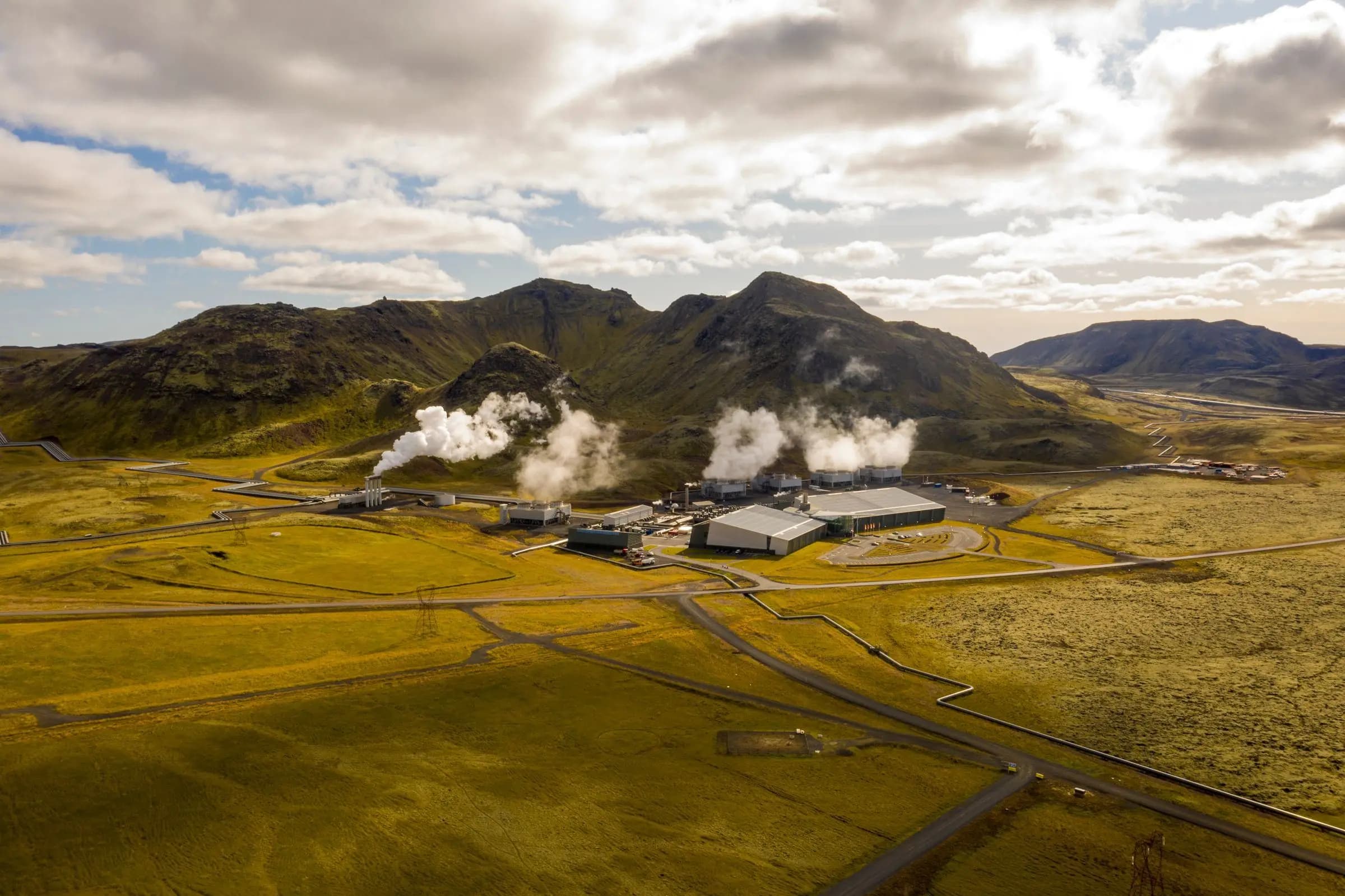 A geothermal power plant venting steam below volcanic ridges, its pipelines crossing open ground